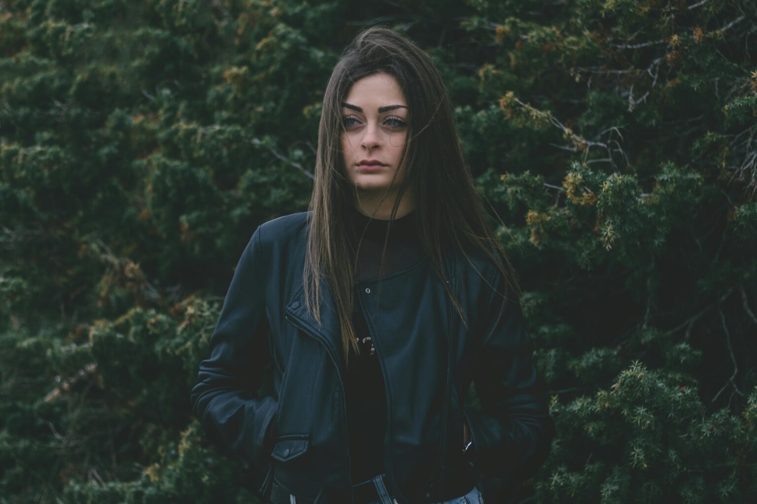 SMM A woman in a leather jacket stands against a lush green backdrop, exuding mystery.