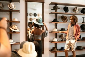 About Us A woman tries on a stylish hat in a boutique store, admired by a companion.