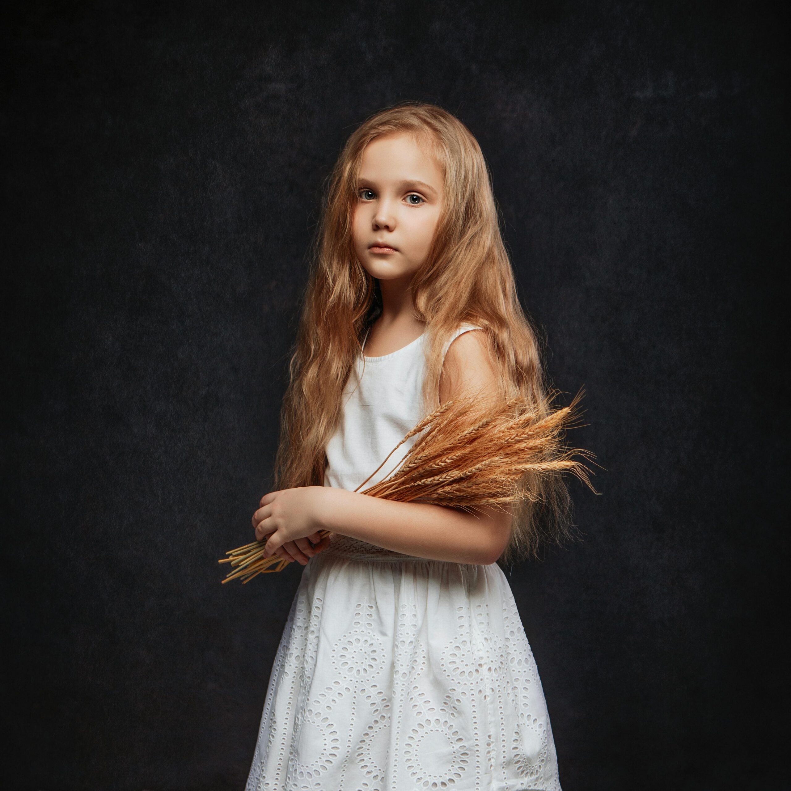 Home A young girl in a white dress holding a wheat bouquet, captured in a classic studio portrait.