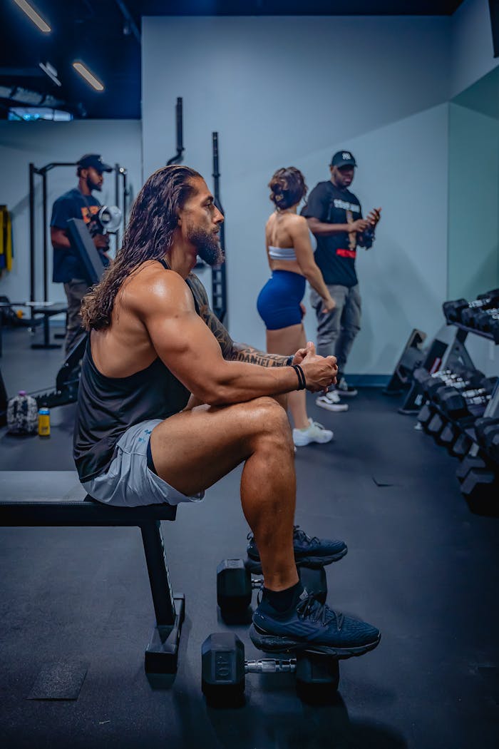 services-02 Muscular man sitting on a weight bench in a modern gym, surrounded by fitness enthusiasts.