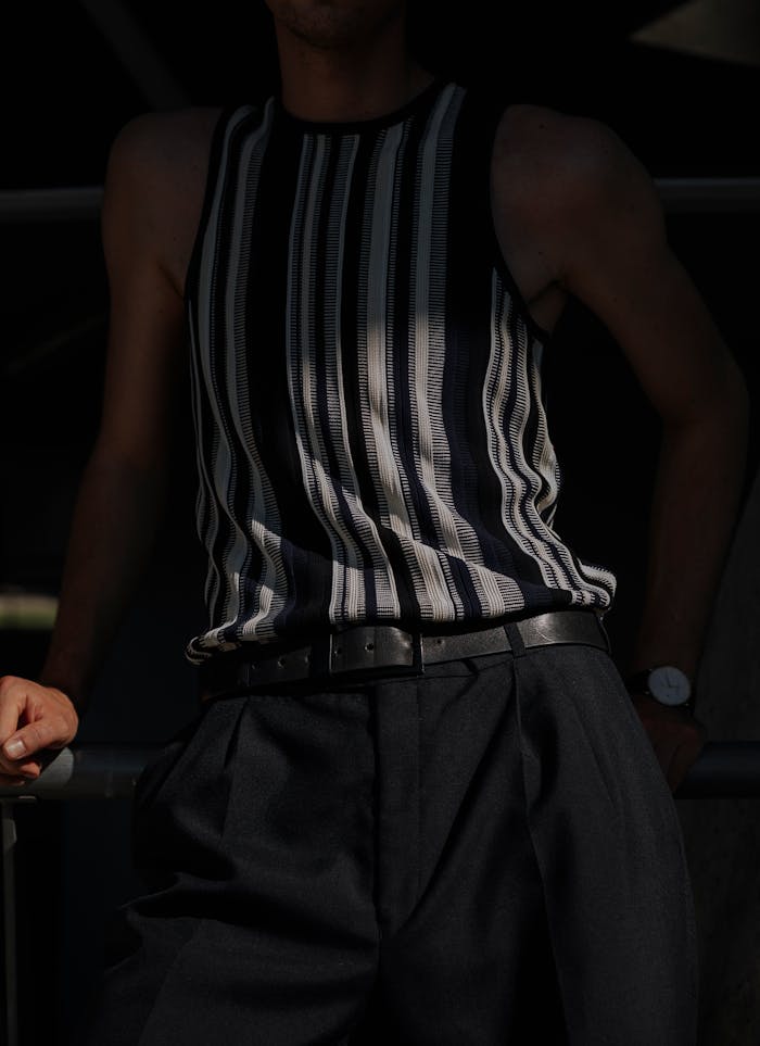 Fashionable man in a striped sleeveless top and dark pants, leaning casually in shadow.